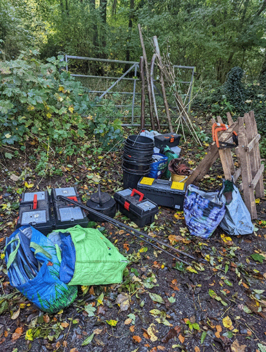 A pile of equipment in front of the gate to the woodlands. Tool boxes, buckets, saws and sawhorses, lots of bags and some wooden sticks.