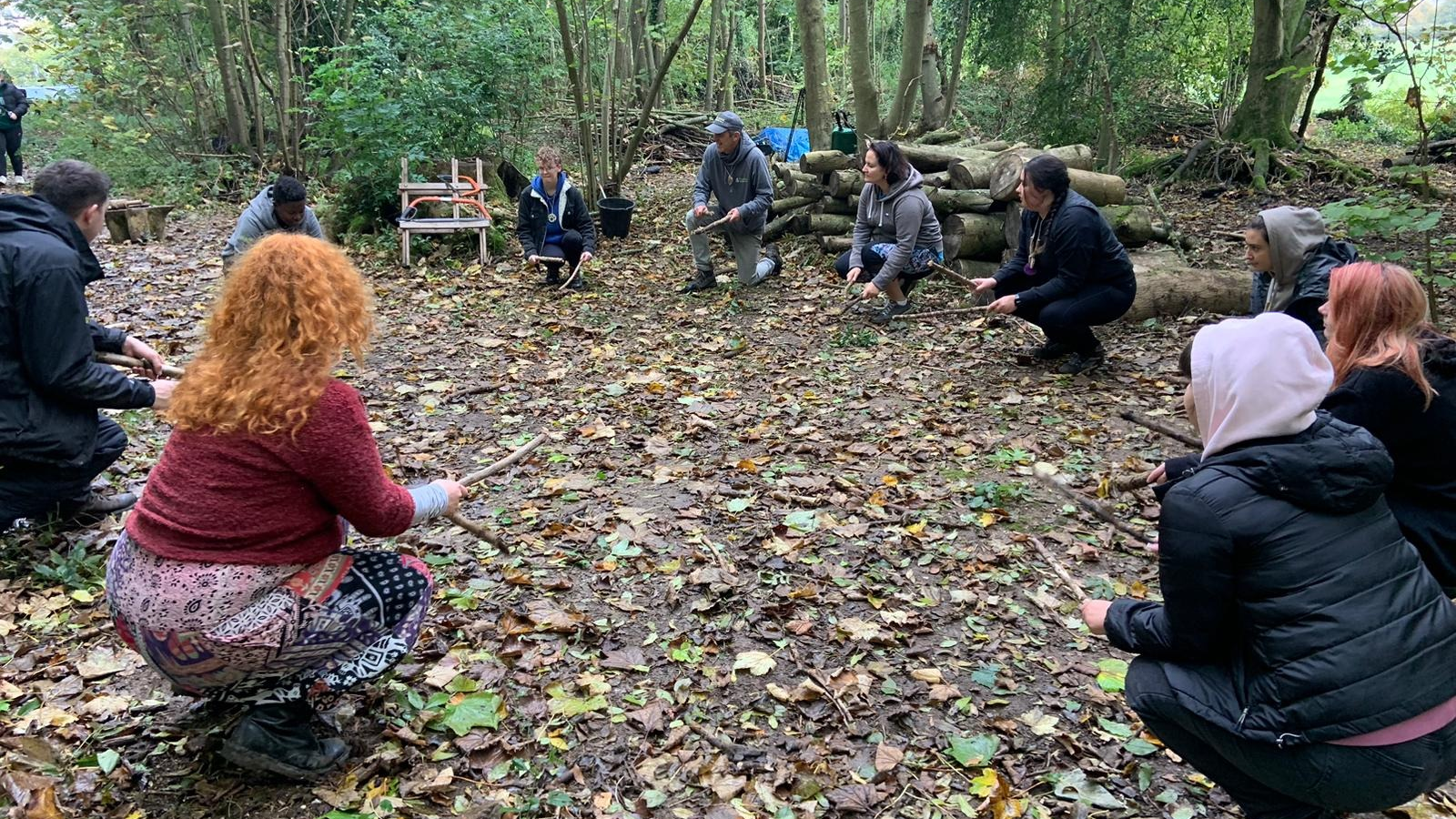 A number of adults in the forest, crouching and holding a stick in each hand.